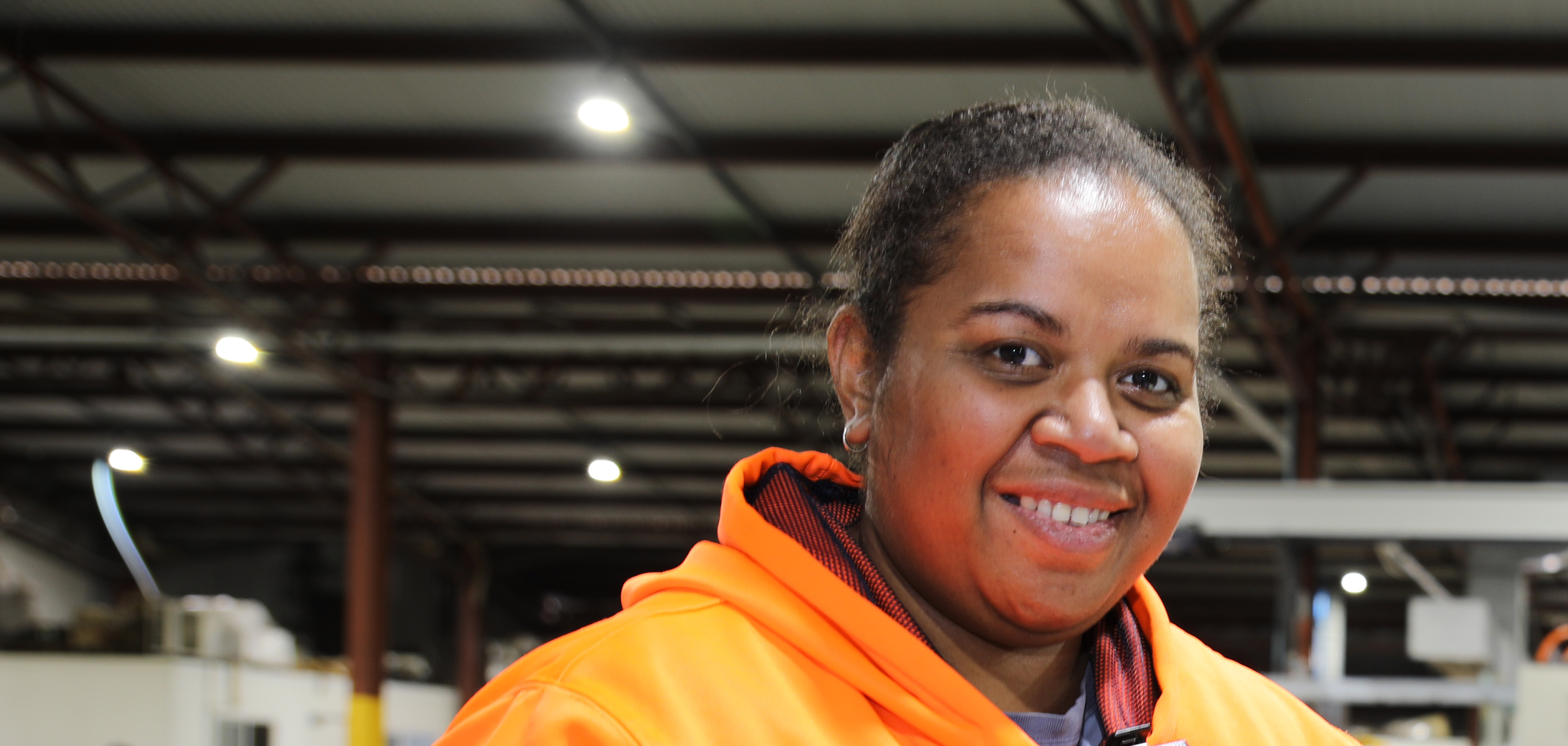 A ni-Vanuatu worker wearing an orange reflective jumper stands in a packing shed. She is smiling and looking at the camera.