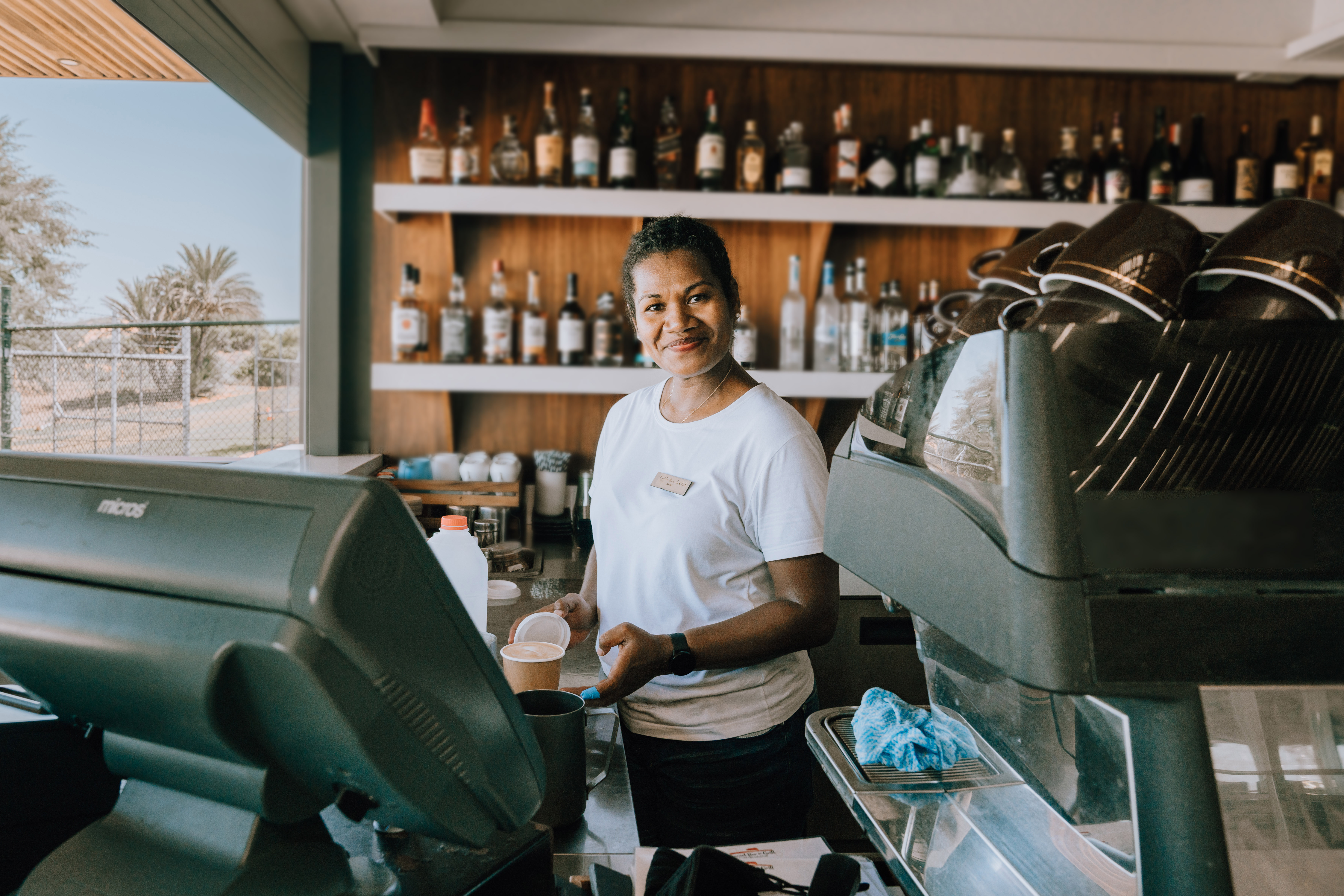 A woman in a white shirt smiles as she makes a coffee at a bar.