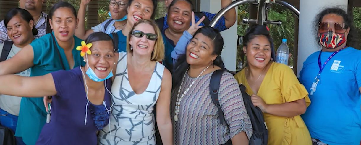 A group of Samoan worker in brightly coloured clothing, some with flowers in the air, stand in a group smiling for the camera with their employer.