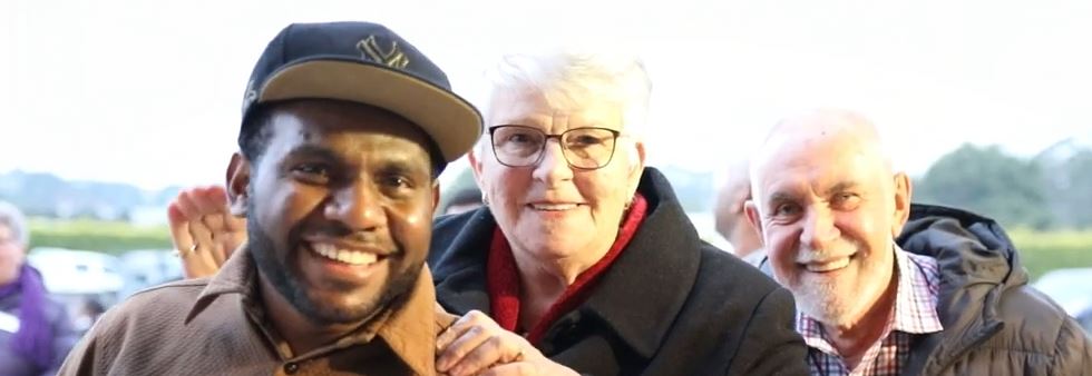 Three people stand together smiling at the camera wearing warm winter clothing. Two are older with grey hair and dark coats, while the man in the foreground has a brown collared shirt and a baseball cap.