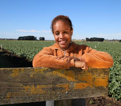 A woman wearing an orange fleecy jumper stands in a field, with blue sky above and a field of lettuces behind her. She is leaning on a wooden gate and smiling at the camera.