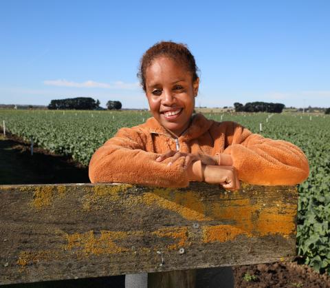 A woman wearing an orange fleecy jumper stands in a field, with blue sky above and a field of lettuces behind her. She is leaning on a wooden gate and smiling at the camera.