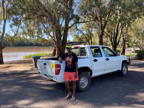 A Ni-Vanuatu woman in a black shirt and pink shorts stands in front of a white ute next to a river.