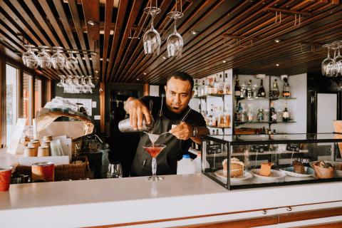 A man in a black shirt and striped apron makes a cocktail at a bar.