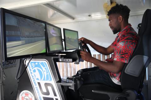A man in a red and black shirt sits at the wheel of a driving simulator.