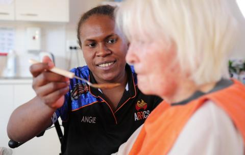 An aged care worker facing the camera holds a spoon up to the mouth of an elderly person.