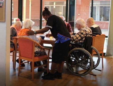 An aged care worker places her hand gently on the back of an elderly person, who is sitting in a circle around a table with four other aged care residents.
