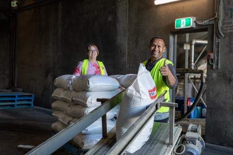 A man and a woman look at the camera smiling, moving sacks on to a conveyer belt