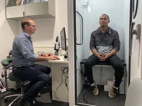 A man sitting in a hearing booth with an audiologist on the other side 