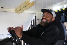 A man in a grey cap and black hoodie sits behind a steering wheel.
