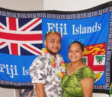 A Fijian man and woman wearing colorful clothing and standing in front of a Fijian flag.