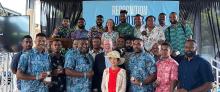 A group of men, mostly wearing colourful printed shirts and holding medals, stand with 3 officials in front of a hanging blue banner that says 'Recognition'