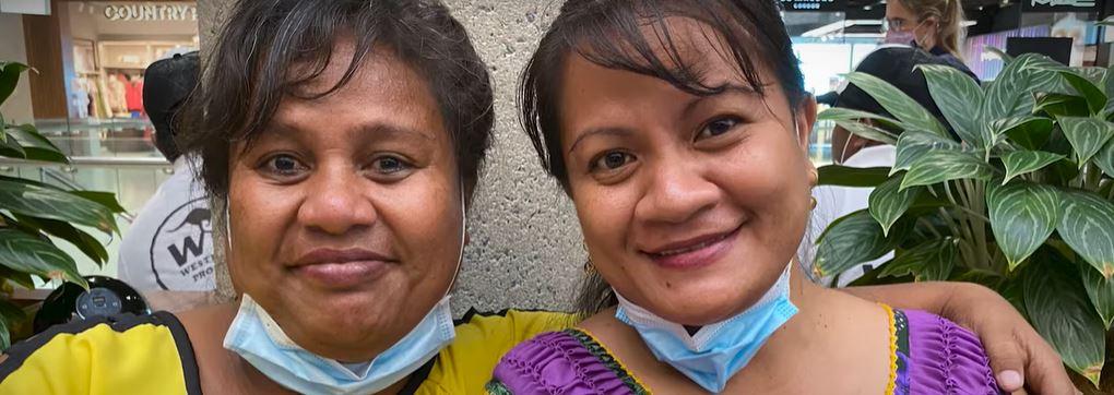 Two Kiribati workers with colourful clothing and with face masks under their chins smile for the camera.