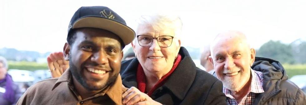 Three people stand together smiling at the camera wearing warm winter clothing. Two are older with grey hair and dark coats, while the man in the foreground has a brown collared shirt and a baseball cap.