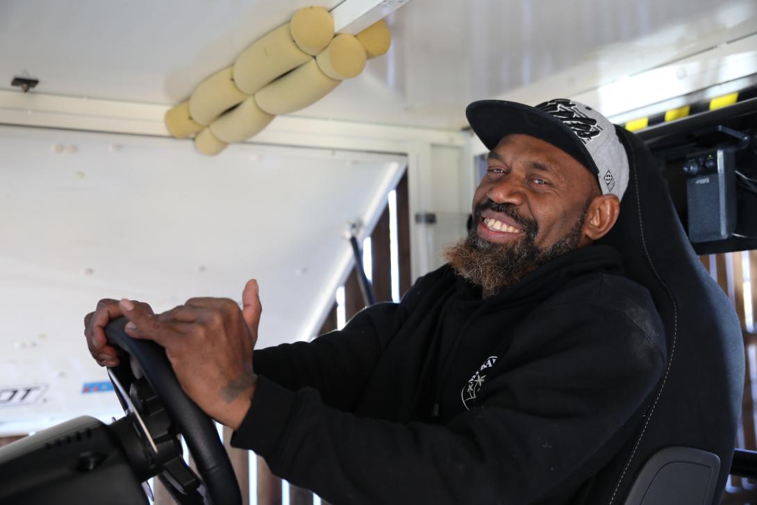 A man in a grey cap and black hoodie sits behind a steering wheel.