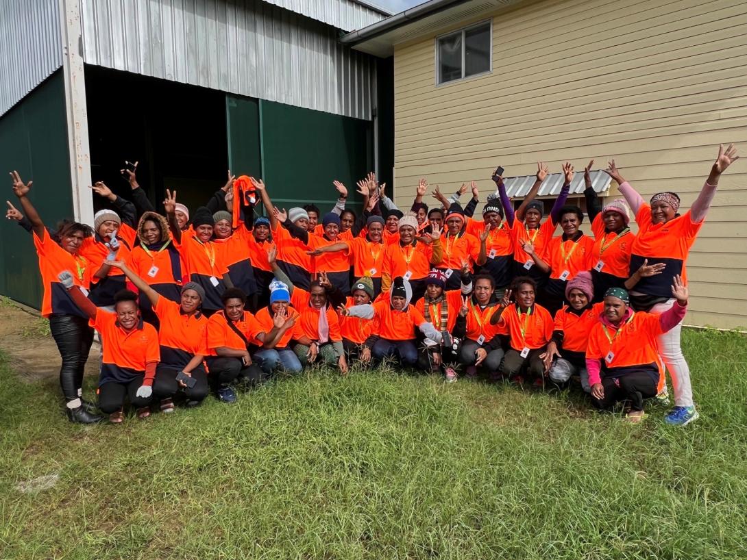 A large group of PNG women in orange high-visibility workwear in front of a shed.