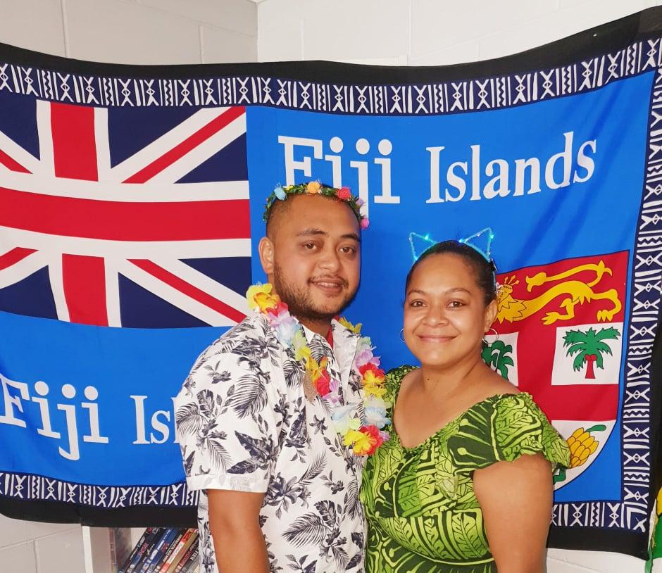 A Fijian man and woman wearing colorful clothing and standing in front of a Fijian flag.
