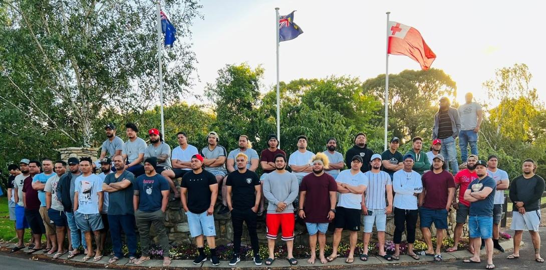 A large group of Tongan men stand in front of flagpoles.