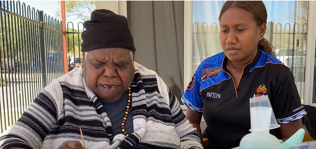 A resident of a personal care facility wearing a black beanie and black and white striped shirt paints a colourful picture, while a residential aged care worker watches.,