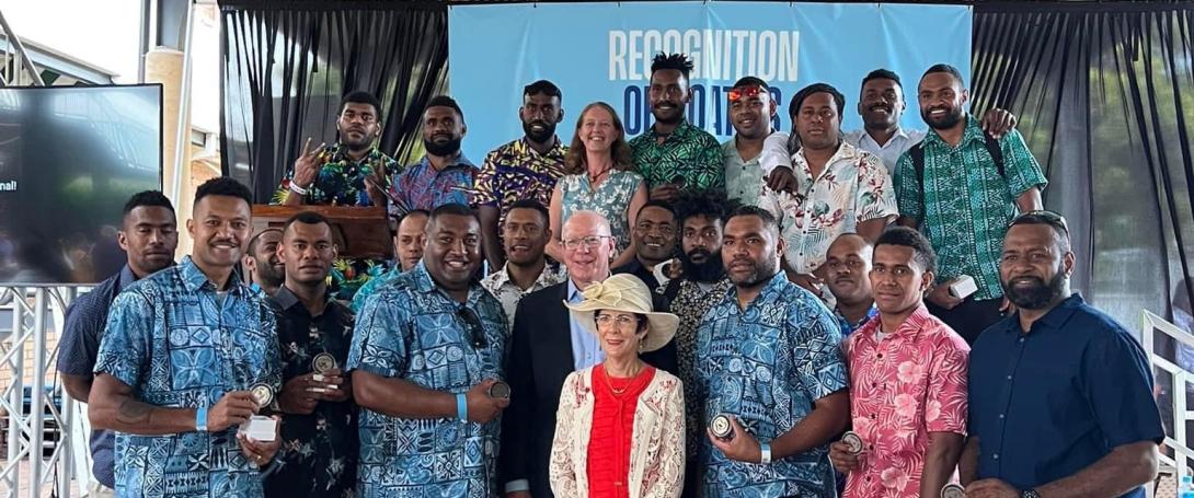A group of men, mostly wearing colourful printed shirts and holding medals, stand with 3 officials in front of a hanging blue banner that says 'Recognition'