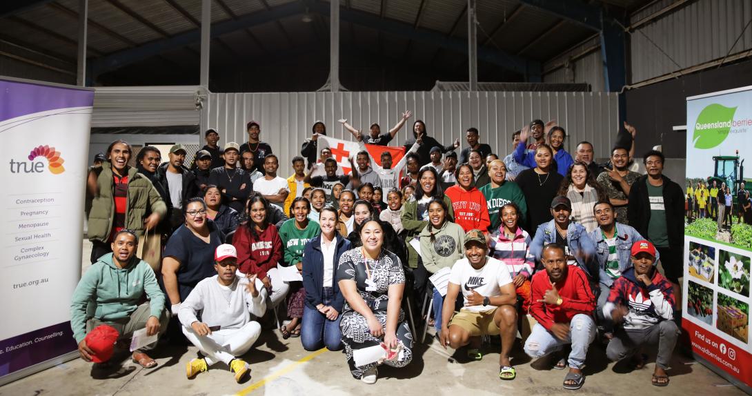 A large group of people smiling at the camera beside a banner. There is a Tongan flag in the background